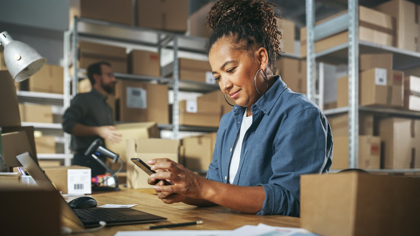 Female business owner in warehouse sitting at her desk, checking her phone while smiling